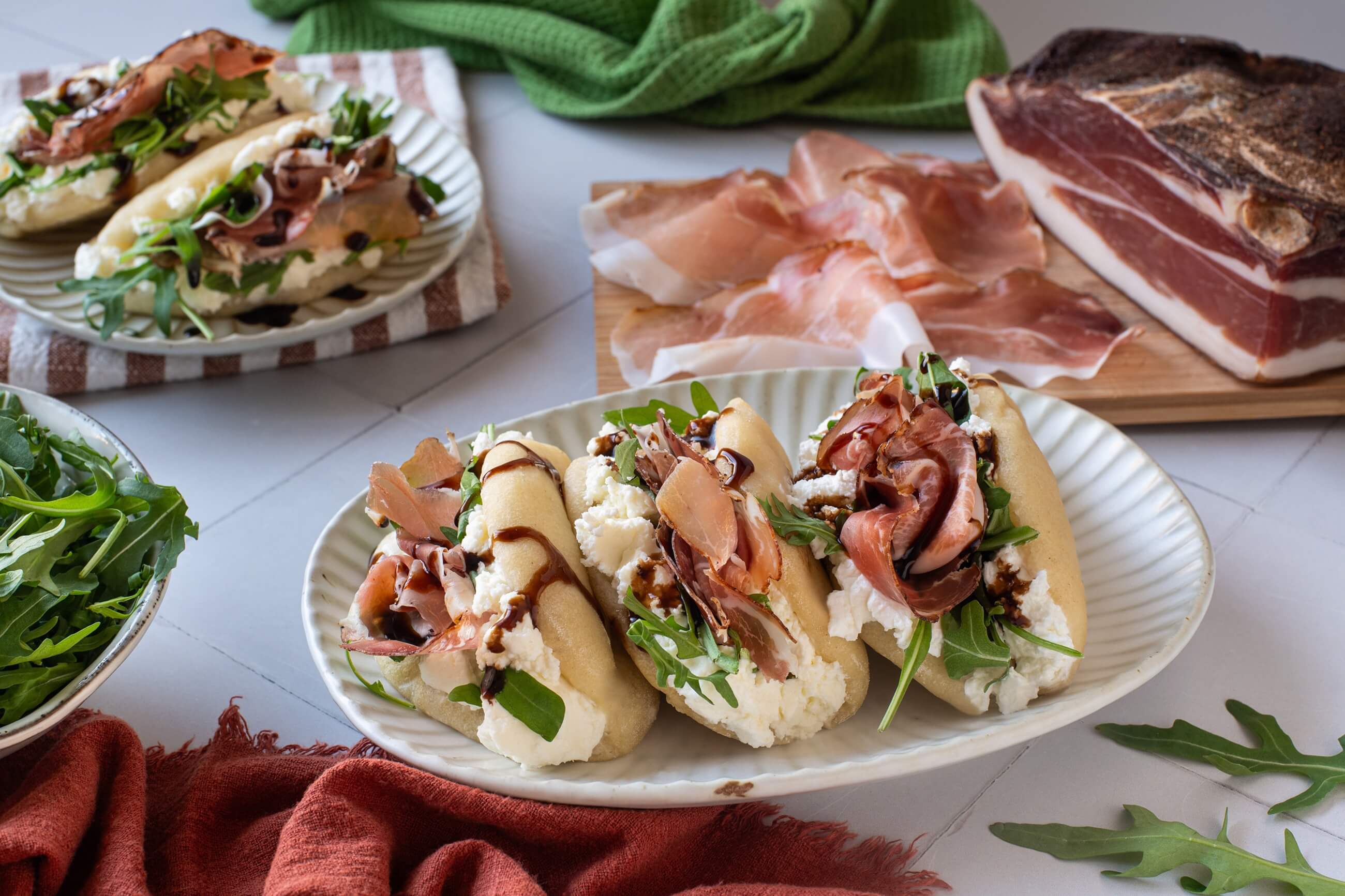 Gedämpfte Bao Buns mit Südtiroler Speck g.g.A., frischer Robiola, Rucola und Balsamicoglasur, serviert auf einem Teller neben aufgeschnittenem Speck.