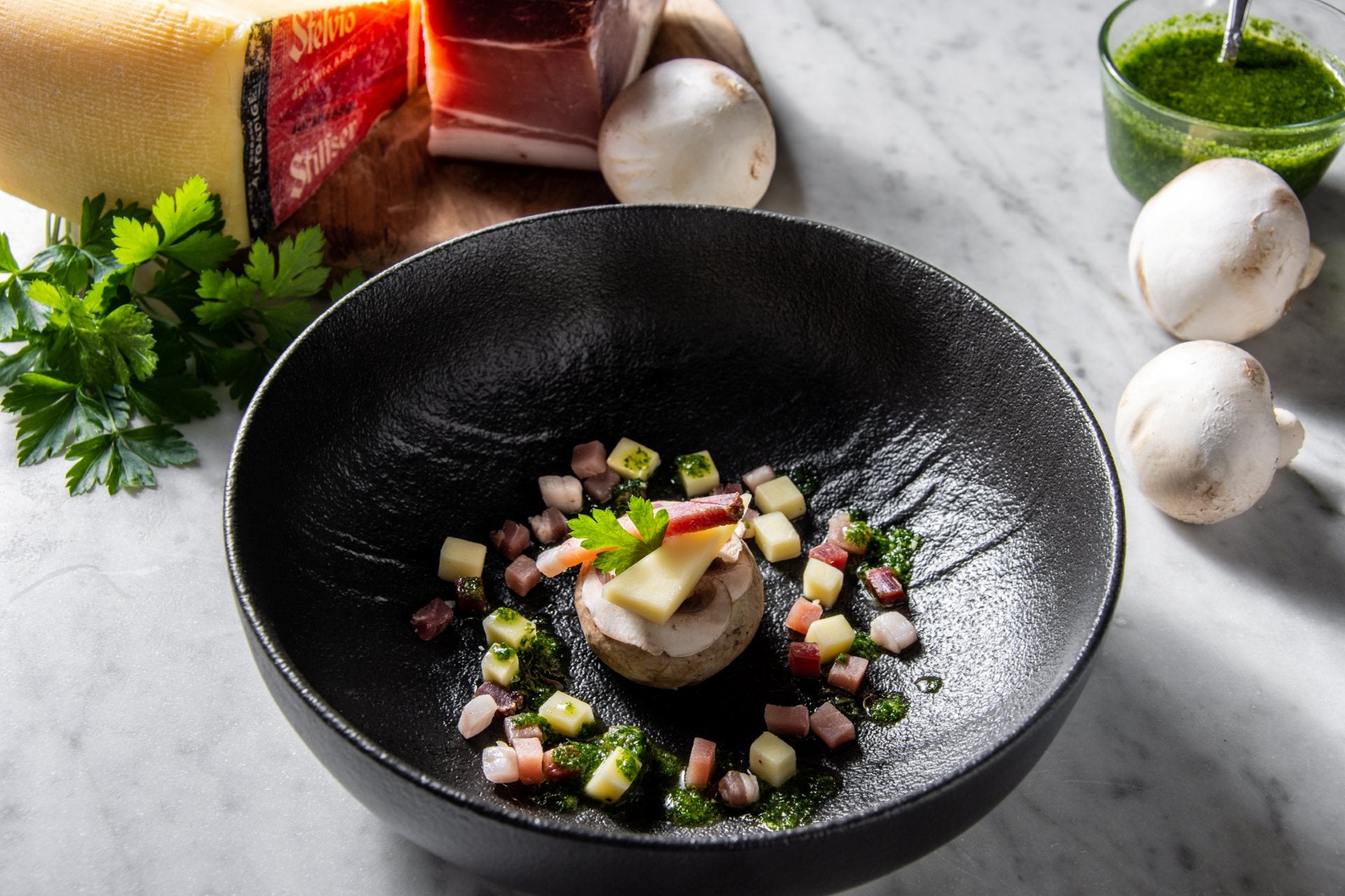 Champignon gefüllt mit Kartoffelcreme, Käse und Südtiroler Speck, angerichtet in einer schwarzen Schale mit Speckwürfeln, Kräutern und grüner Sauce.