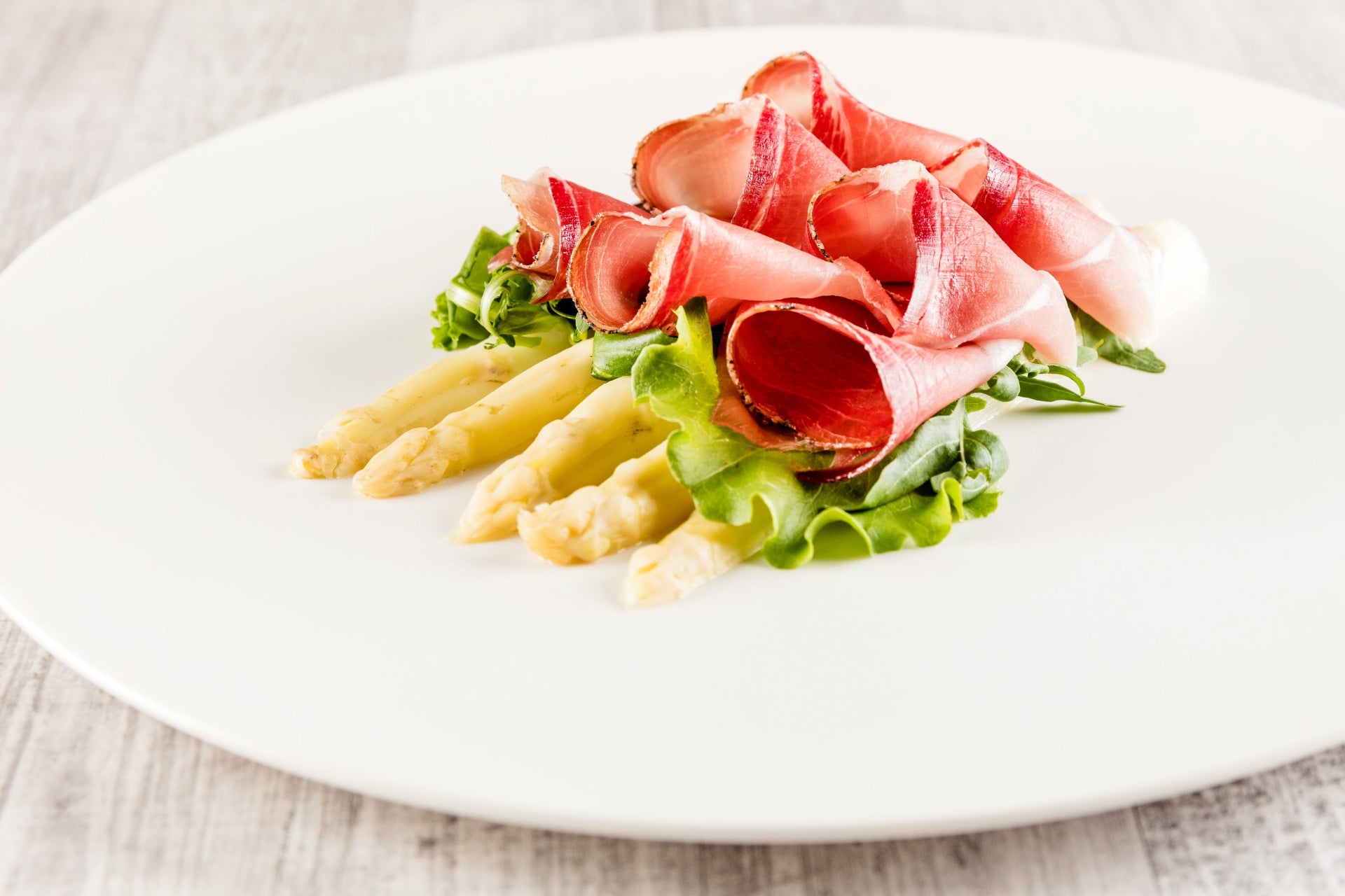 Spargelsalat mit Carpaccio vom Südtiroler Speck g.g.A. und Blattsalat, elegant angerichtet auf einem weißen Teller.