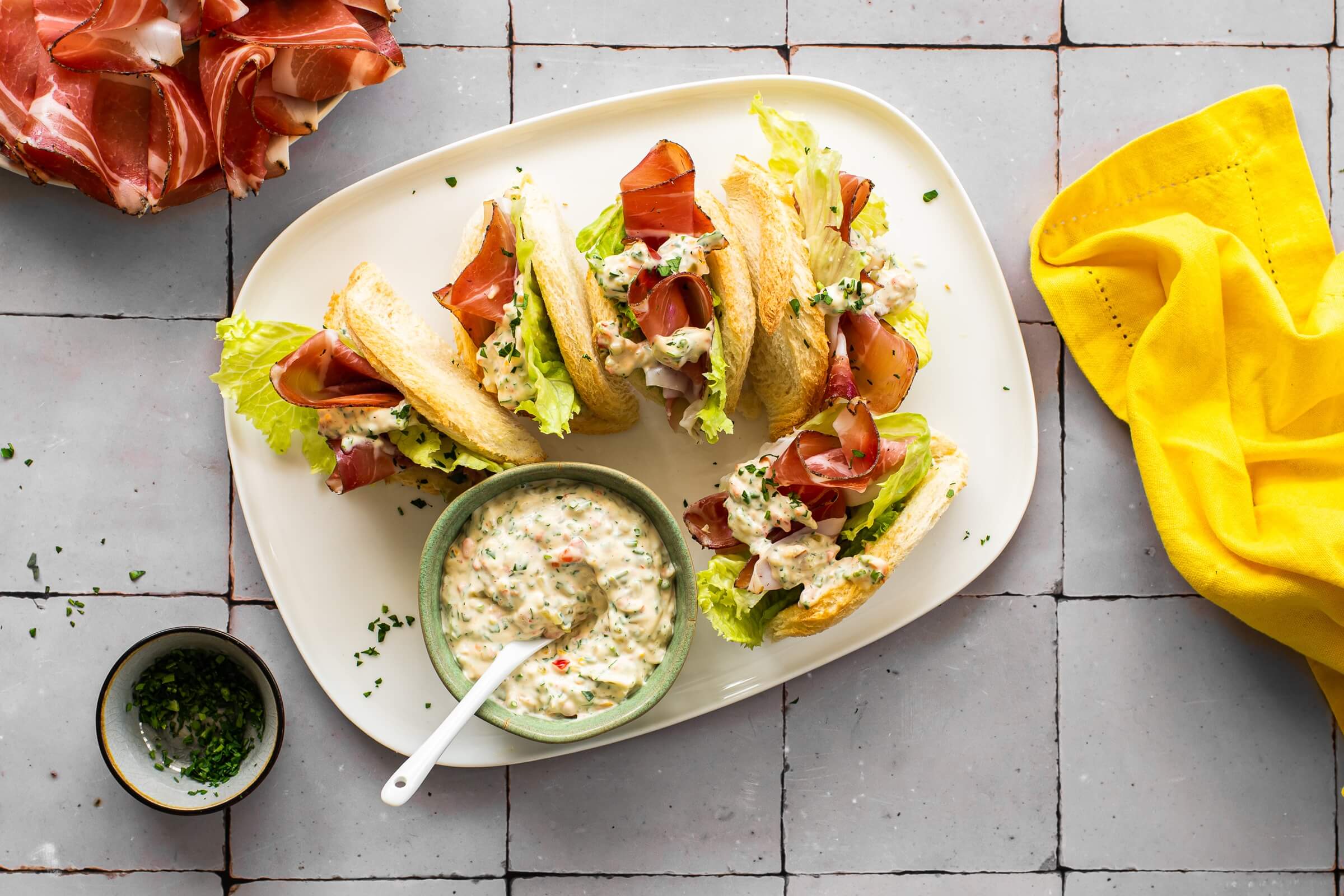 Offen belegte kleine Sandwiches auf einem weißen Teller, gefüllt mit Salatblättern, Tartarsauce und Scheiben von Südtiroler Speck g.g.A.; in der Mitte des Tellers steht eine kleine Schüssel mit derselben Creme und einem Teelöffel. Im Hintergrund sieht man weitere Scheiben von Südtiroler Speck g.g.A. und ein gelbes Küchentuch.