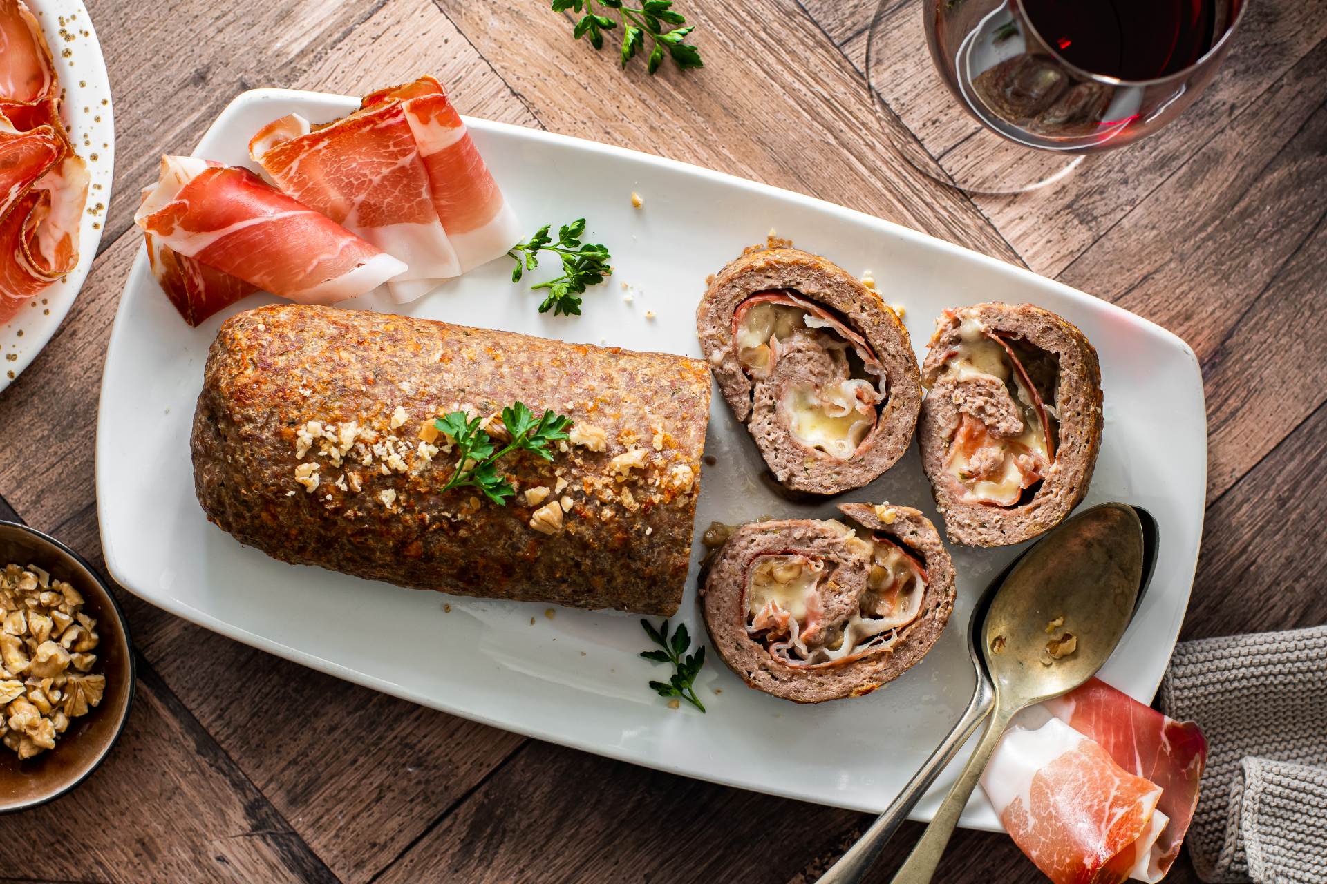 Gefüllter Hackbraten mit Südtiroler Speck und Käse, in Scheiben geschnitten auf einer weißen Platte serviert, mit gehackten Nüssen und Petersilie garniert, dazu Speck und ein Glas Rotwein.