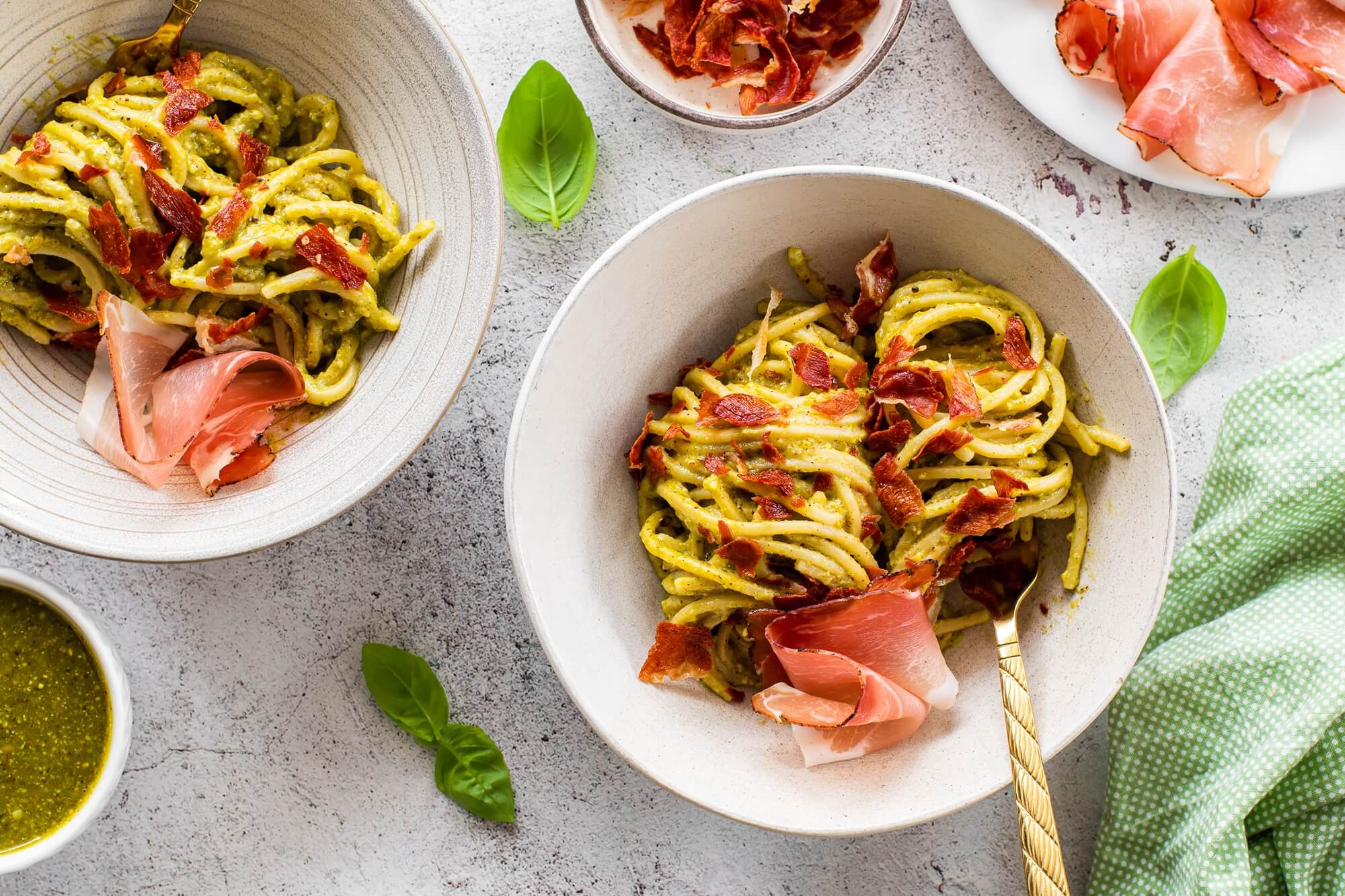 Schüssel mit Spaghetti, daneben Pesto, Basilikumblätter und Speckchips.