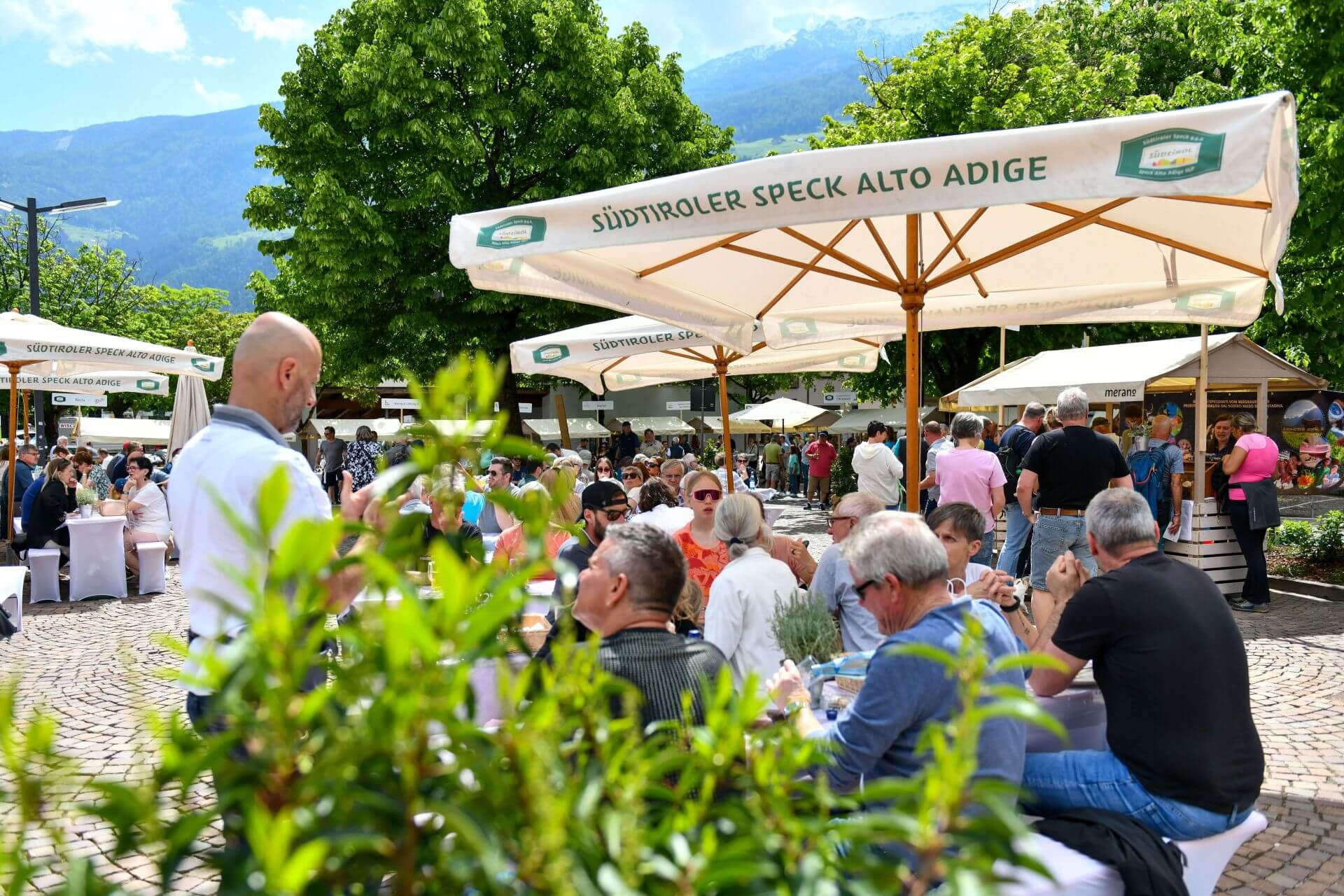 Menschen sitzen an Tischen bei Veranstaltung im Freien unter Sonnenschirmen mit Speck Alto Adige g.g.A. Schriftzug