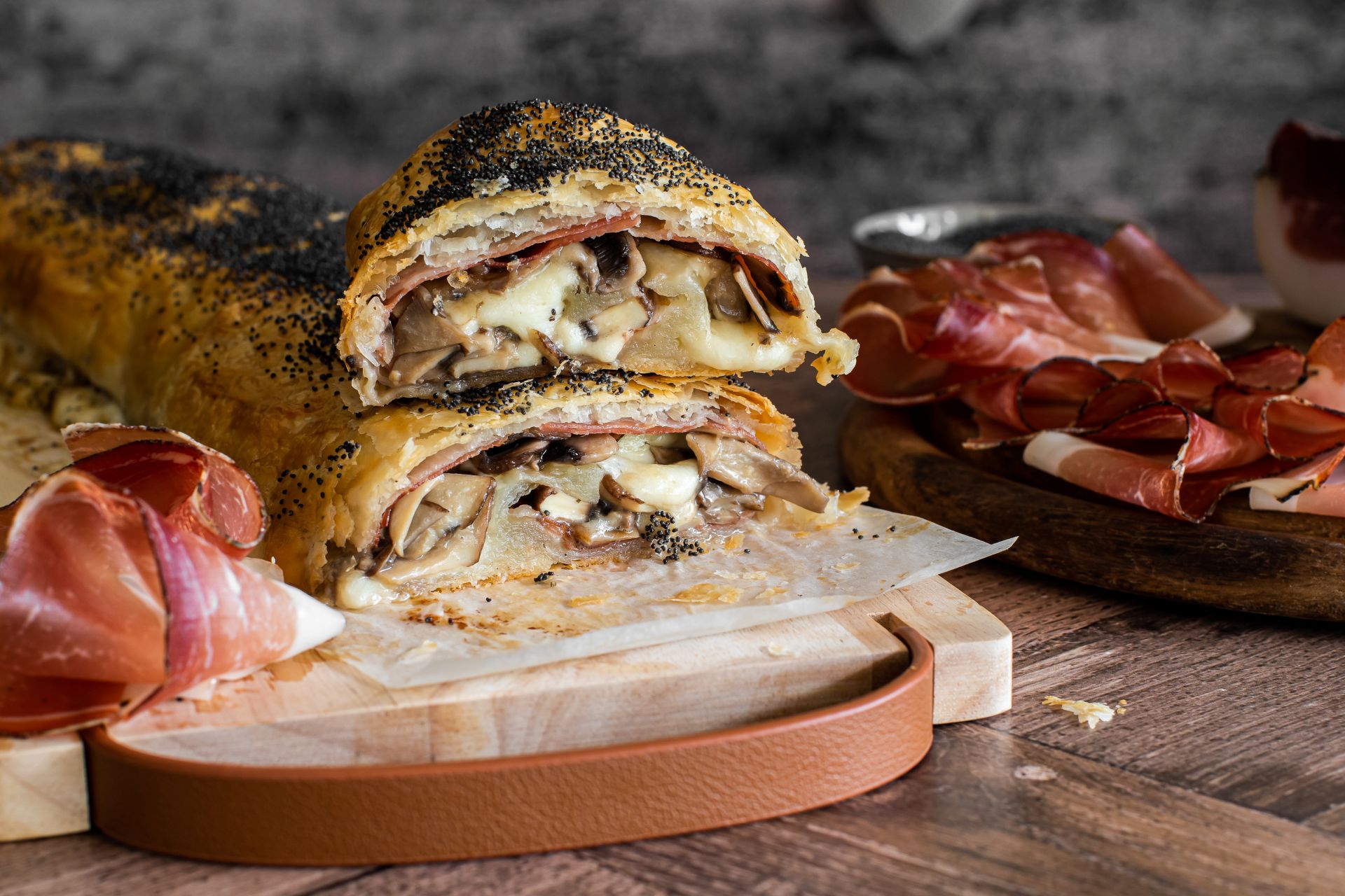 Herzhaftes Strudel aufgeschnitten mit einer Füllung aus Champignons, Südtiroler Speck g.g.A. und geschmolzenem Käse, serviert auf einem Holzbrett mit Speckscheiben daneben.