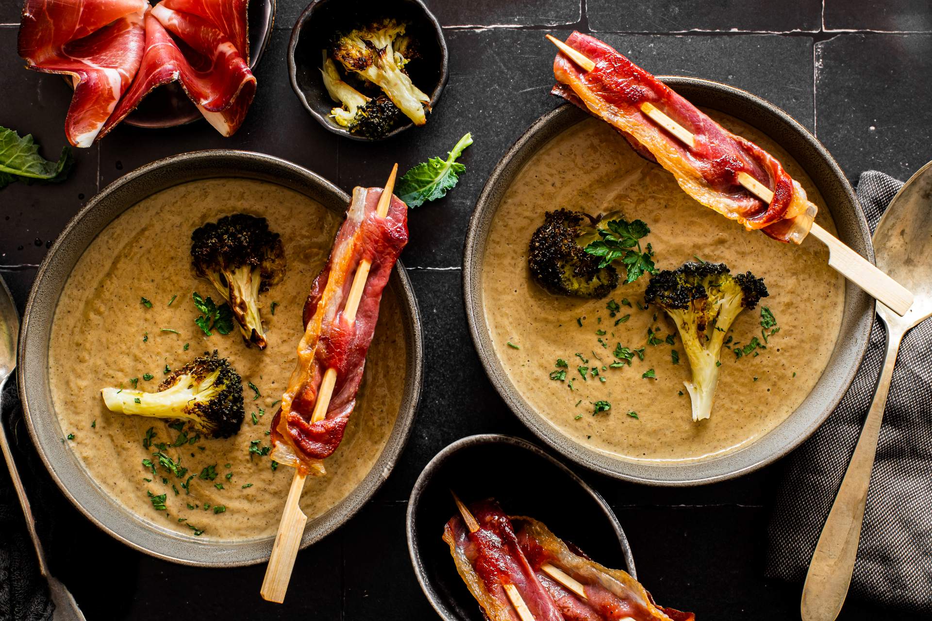 Brokkolicremesuppe in einer Schale, garniert mit gerösteten Brokkoliröschen und einem Spießchen vom Südtiroler Speck g.g.A., mit frischen Kräutern bestreut.