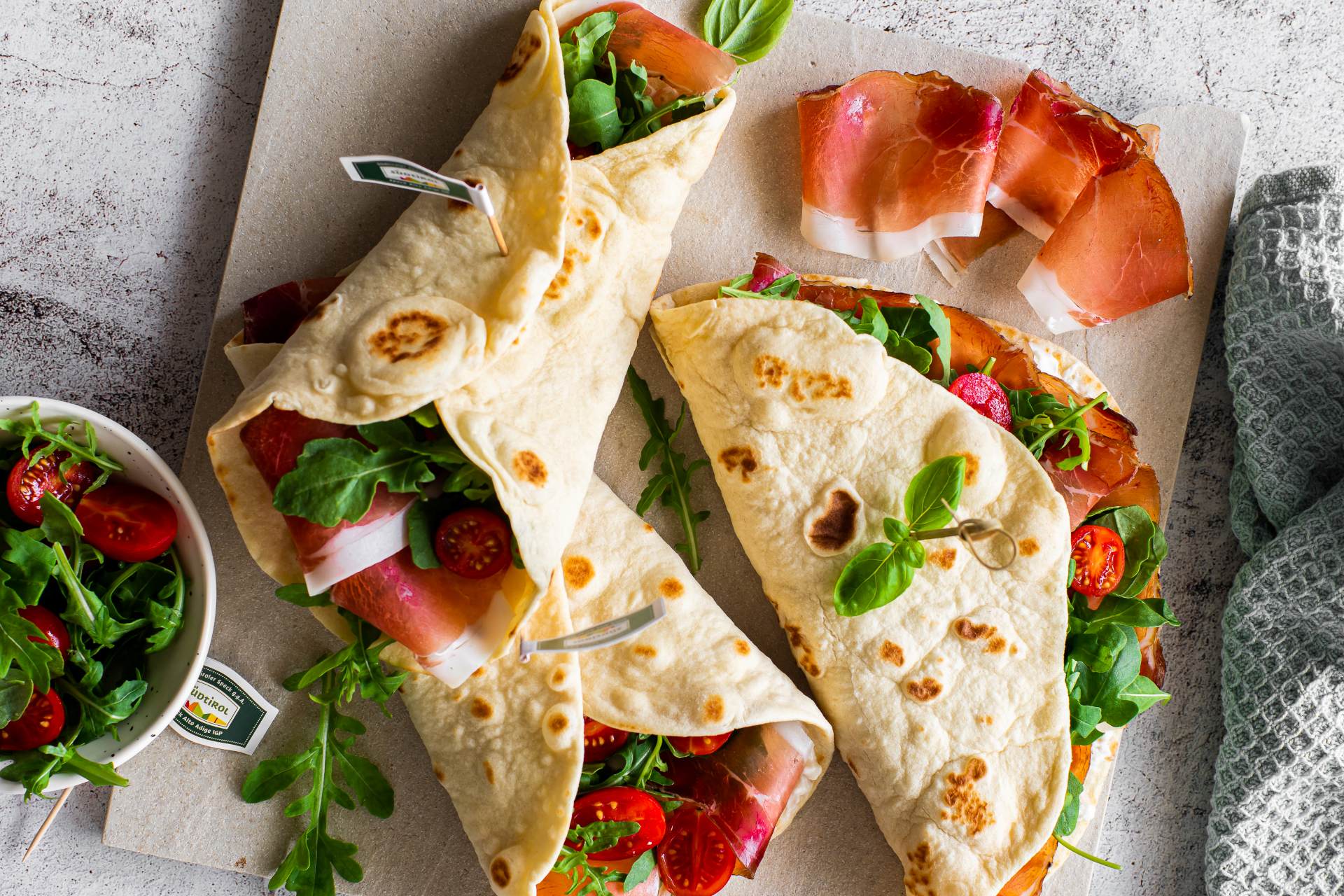 Gefaltete Piadine, gefüllt mit Südtiroler Speck g.g.A., Rucola und Kirschtomaten, angerichtet auf einem Schneidebrett, mit Speckstreifen daneben und einer Schüssel Salat.