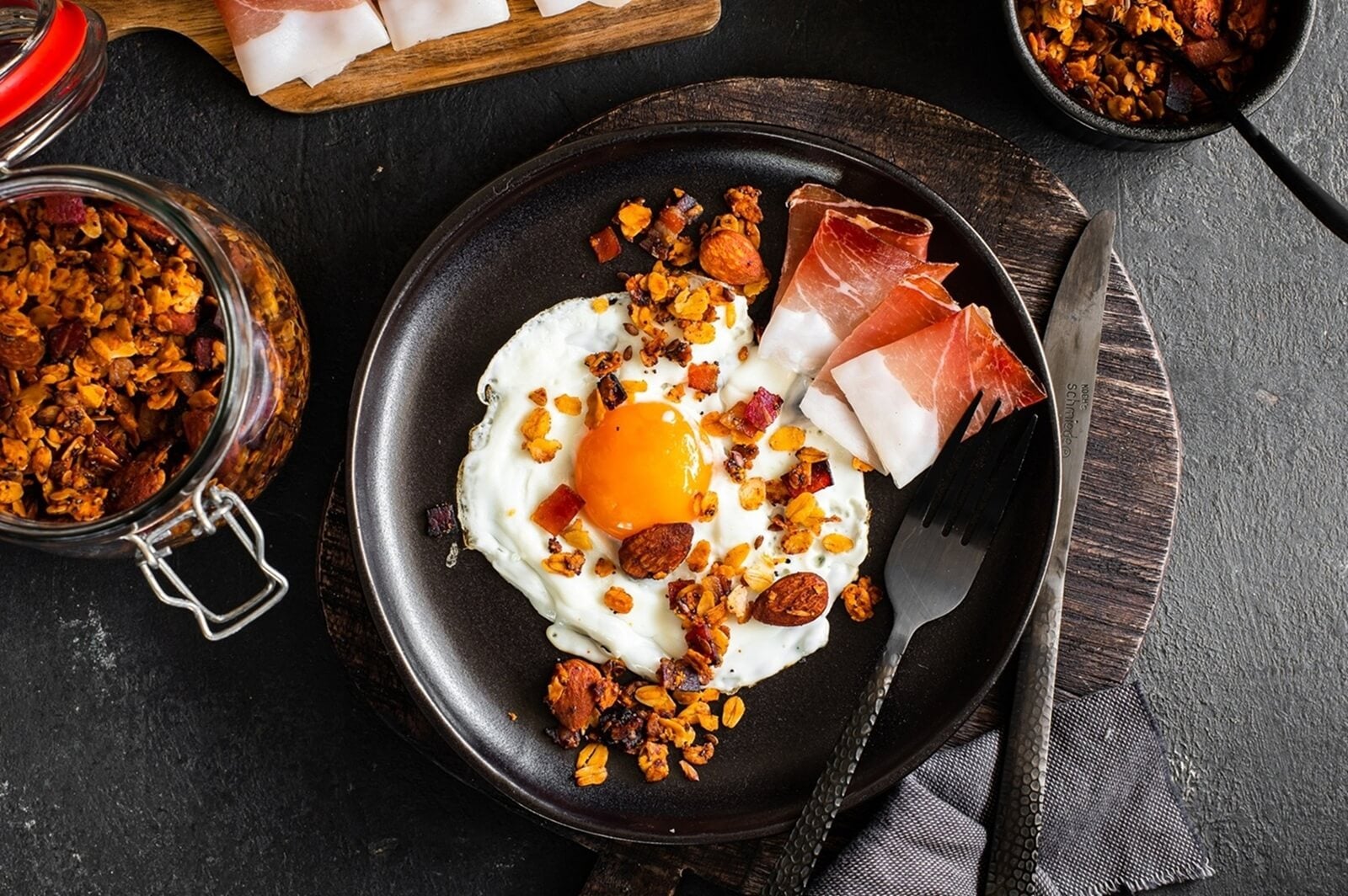 Spiegelei mit weichem Dotter auf dunklem Teller serviert, begleitet von knusprigem Granola und dünnen Scheiben Südtiroler Speck g.g.A., mit Edelstahlbesteck und einem Südtiroler Speck g.g.A Schneidebrett im Hintergrund.