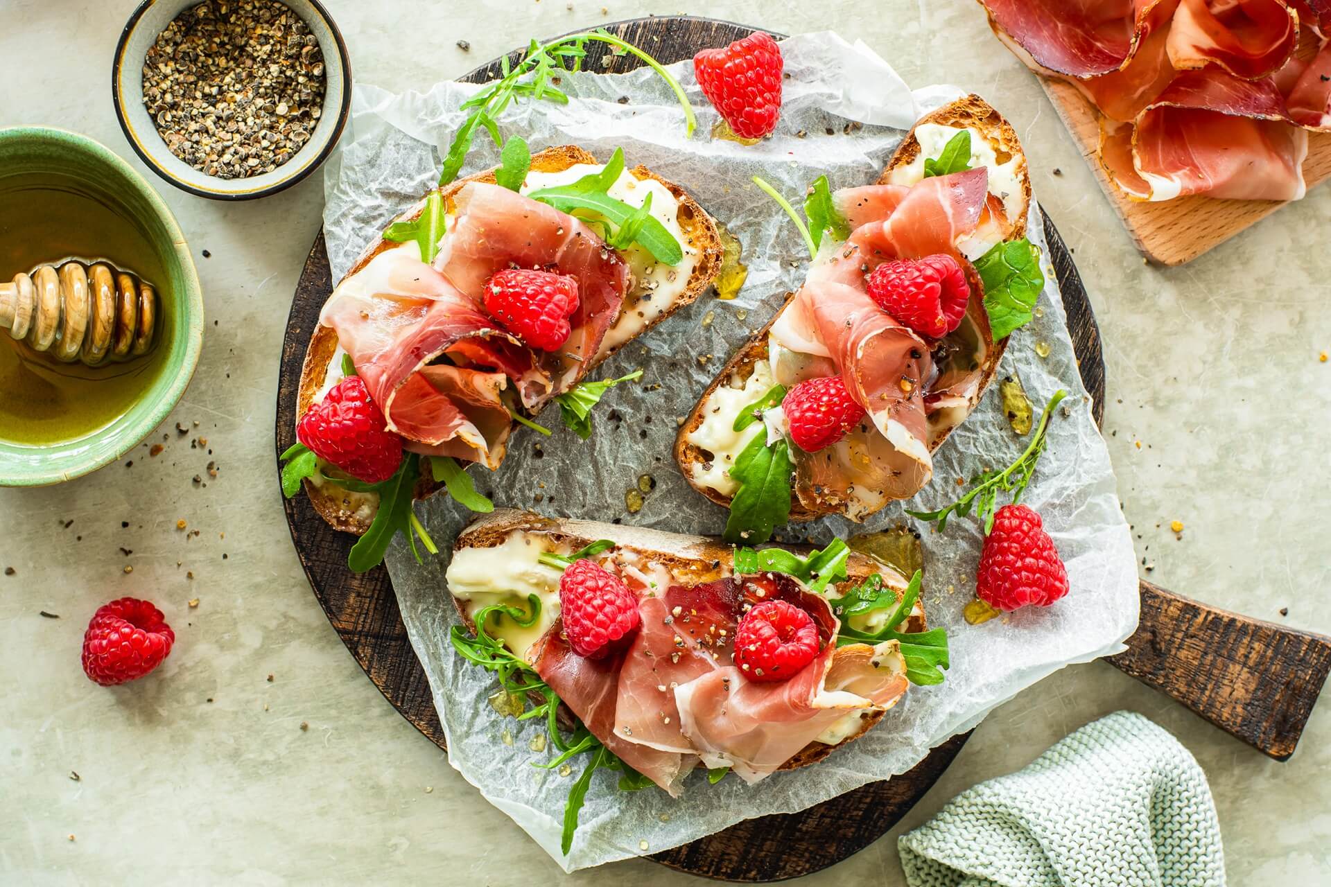 Knusprige Crostini mit Gorgonzola, Südtiroler Speck g.g.A., Rucola und frischen Himbeeren, serviert auf einem Holzbrett mit Honig und Pfeffer.