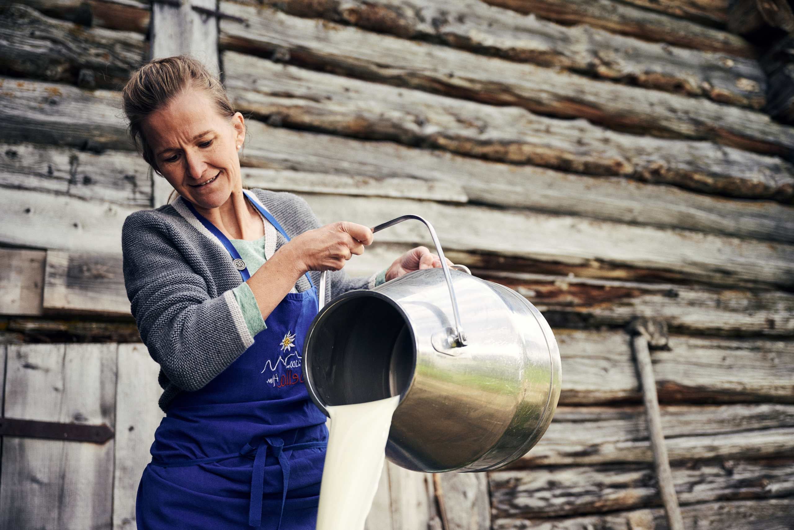 Bäuerin gießt frische Milch aus Kanne vor traditioneller Holzhütte in Südtirol.