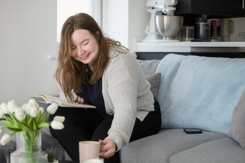 Paige placing her coffee on the table whilst reading a book on the sofa