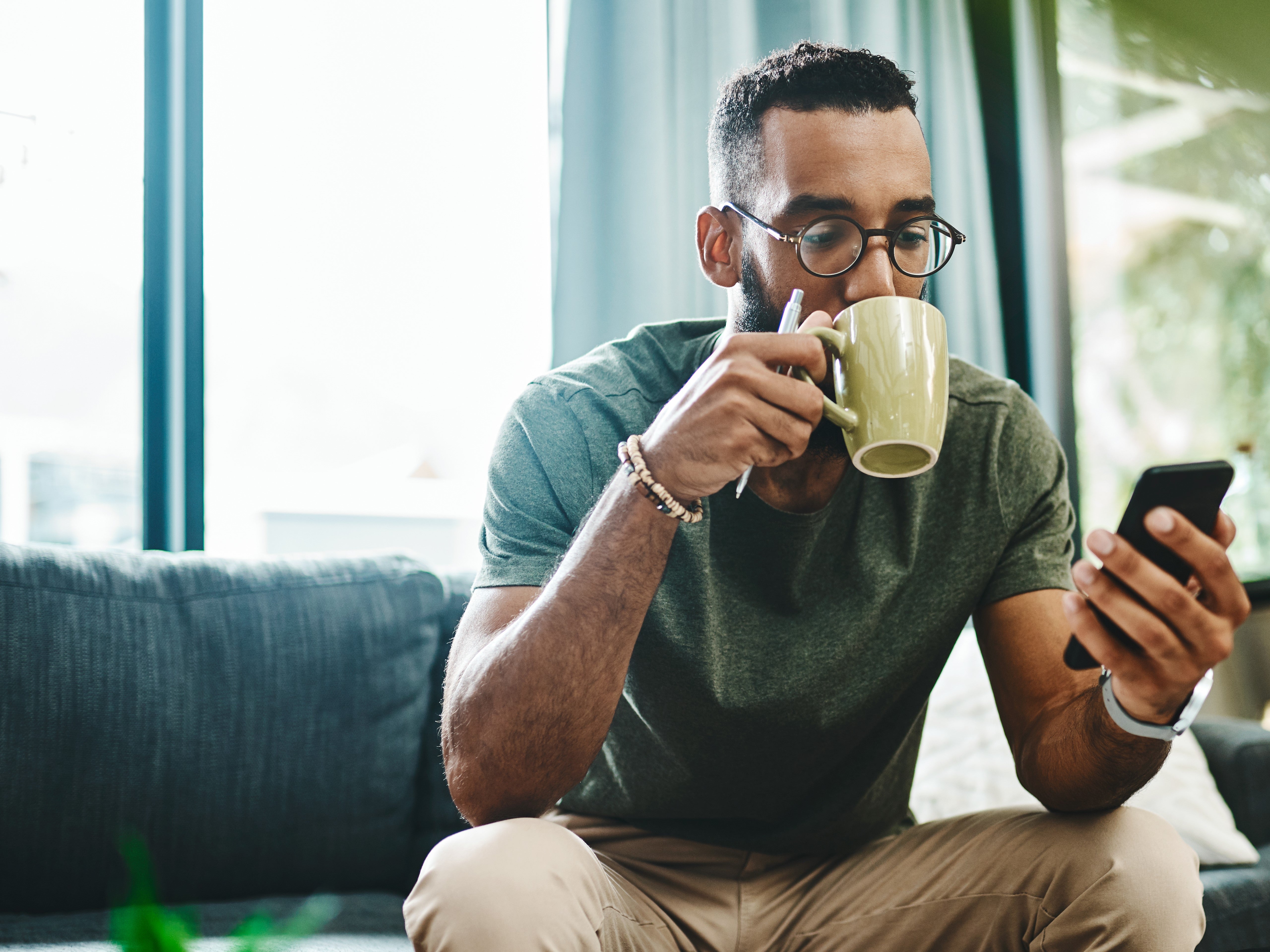 A man sits on a sofa browsing the internet on his mobile phone whilst holding a coffee cup and pen in the other hand.