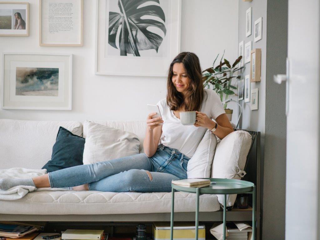 A lady relaxing on the sofa using her phone in one hand and holding a hot drink in the other