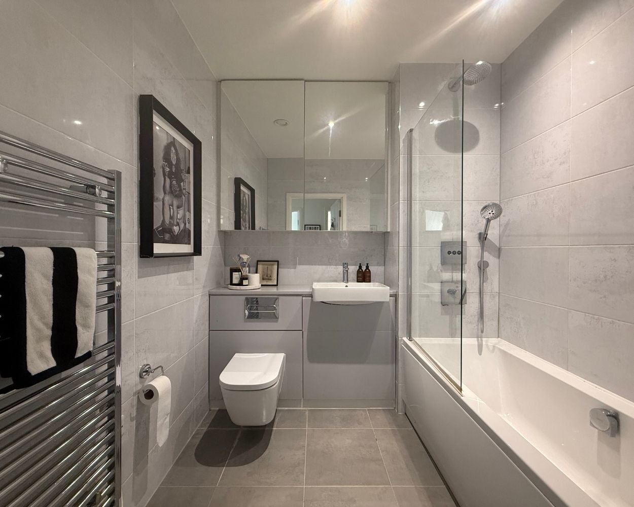 Photo of the grey tiled bathroom, featuring a shower over the bath, large mirrored cabinet and chrome towel rail
