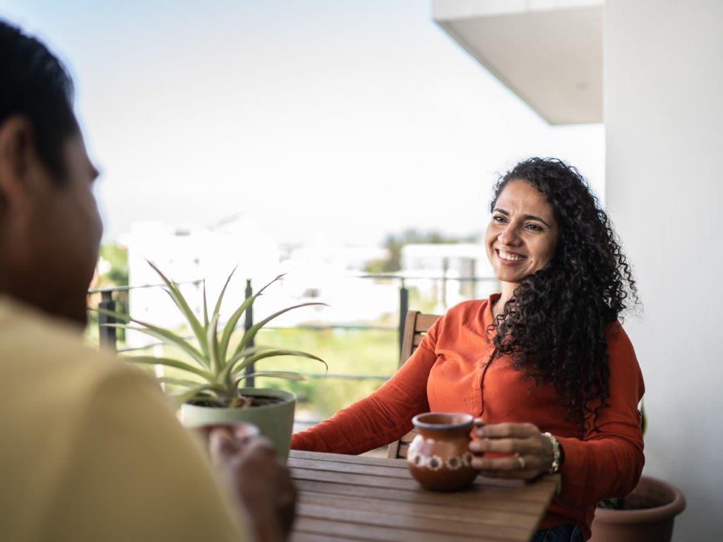 A lady smiles as she has a conversation with a friend whilst sitting at the table on her balcony