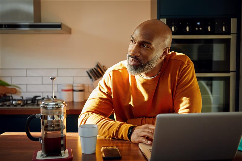 Man stares out of his kitchen window whilst working on his laptop at the breakfast bar.