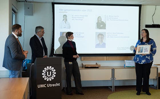 Vier mensen staan voor een scherm tijdens een presentatie bij het UMC Utrecht, waarbij een vrouw een certificaat ontvangt en de anderen toekijken