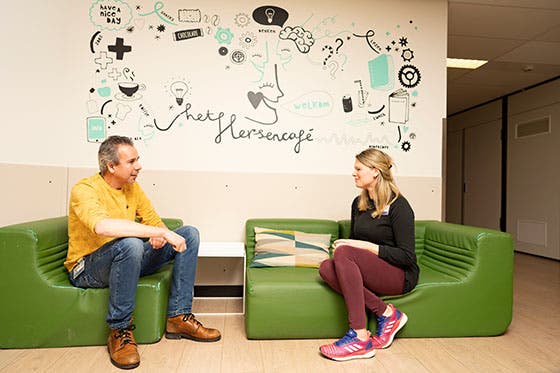 Twee mensen zitten op groene banken in het Hersencafé van het UMC Utrecht, met een muurschildering op de achtergrond die wetenschappelijke thema's weergeeft