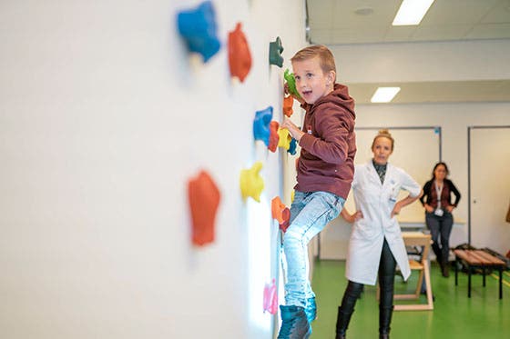 Een jongen klimt op een muur tijdens een revalidatie-oefening, terwijl een arts en een andere begeleider toekijken in een medische faciliteit