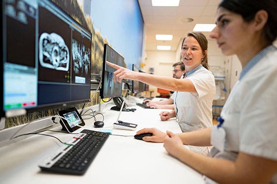 Twee medisch specialisten analyseren MRI-beelden op computerschermen in een controlekamer, waarbij één van hen iets aanwijst op het scherm