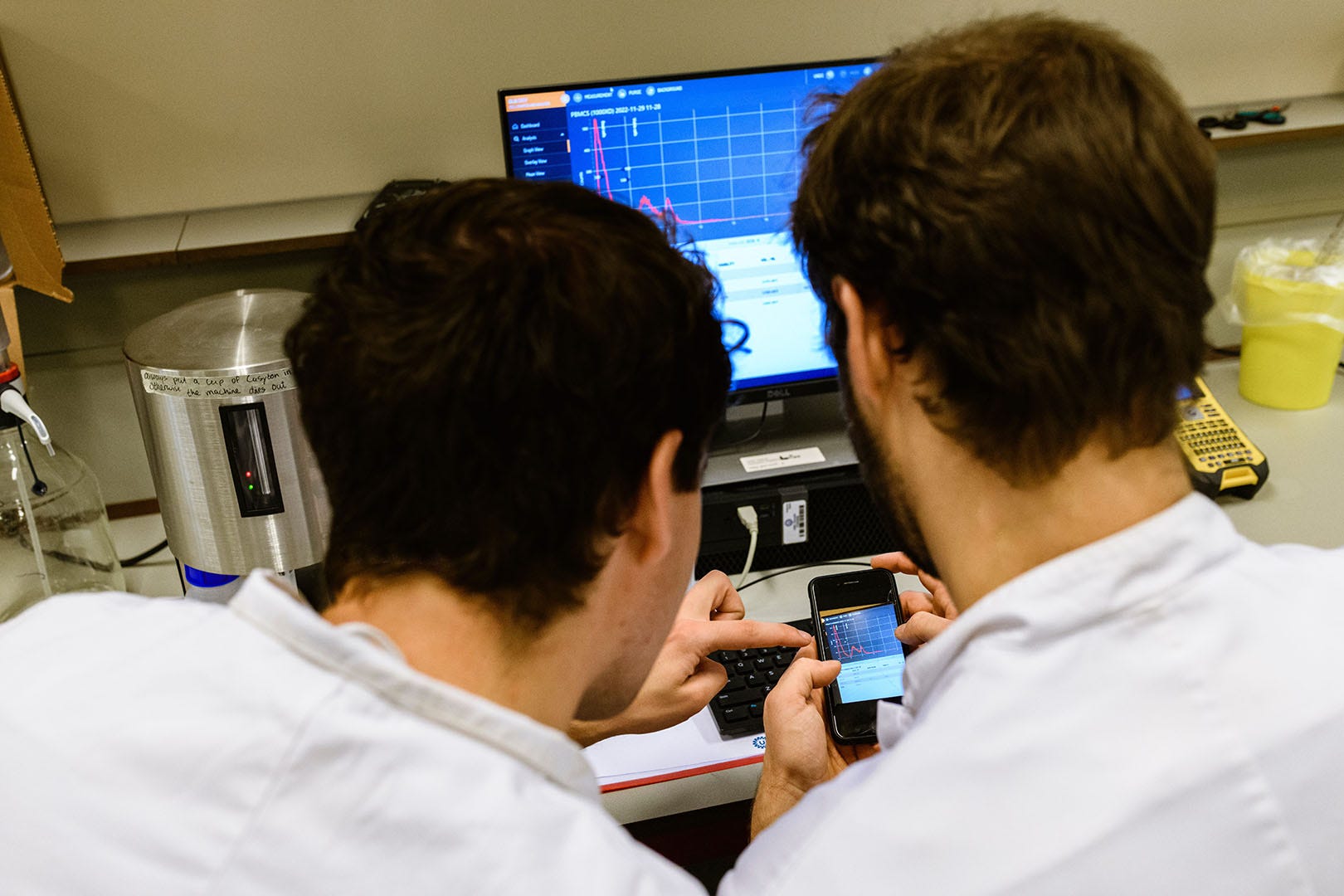 Two scientists in lab coats analyzing data on a smartphone and a computer screen displaying a graph in a laboratory setting