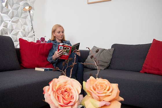 Een vrouw zit op een grijze bank met rode kussens in het gastenverblijf van het UMC Utrecht rustig een boek lezend. Bloemen staan op de voorgrond.