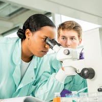Een onderzoeker kijkt door een microscoop terwijl een andere onderzoeker ernaast staat.