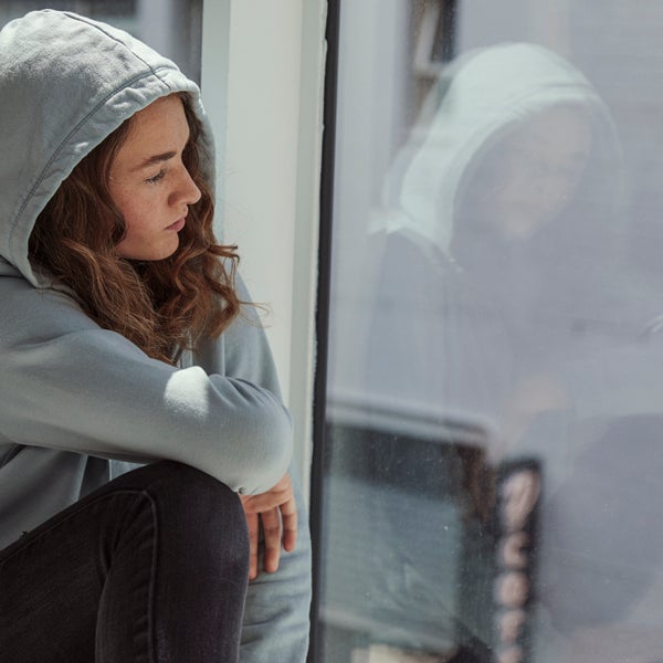 Een jonge vrouw in een grijze hoodie kijkt uit het raam.
