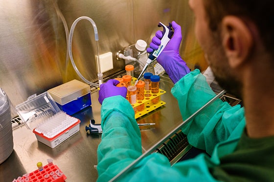 A researcher in a green lab coat and purple gloves uses a pipette to work with test tubes in a laboratory setting.