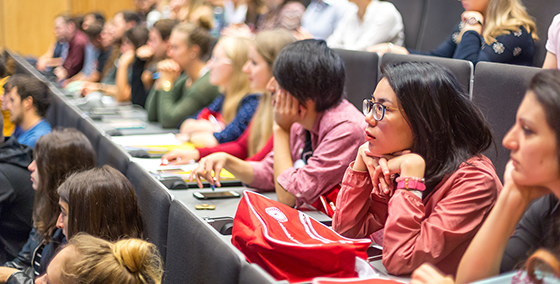Studenten zitten in een collegezaal, aandachtig luisterend naar een lezing. Een studente op de voorgrond draagt een roze blouse en bril.