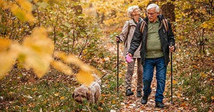 Ouder stel wandelt met hun hond in een bosrijk gebied in de herfst, beiden gebruiken wandelstokken en genieten van de natuur