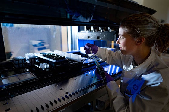 Een laboratoriummedewerker in een witte jas en paarse handschoenen werkt met een pipet bij een high-tech laboratoriumapparaat