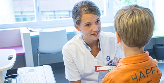 A nurse in a white uniform uses a pain scale to assess a young boy in an orange "HAPPY" shirt, in a bright room with large windows.