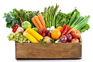 Een houten krat gevuld met diverse verse groenten en fruit, waaronder wortels, asperges, appels, sinaasappels, en maïs, tegen een witte achtergrond