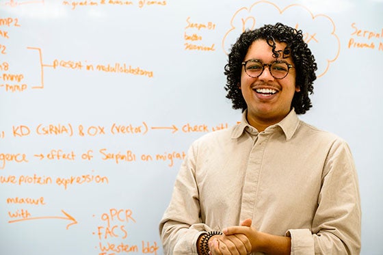 Een lachende student staat voor een whiteboard vol met wetenschappelijke notities en diagrammen tijdens een geneeskundeopleiding