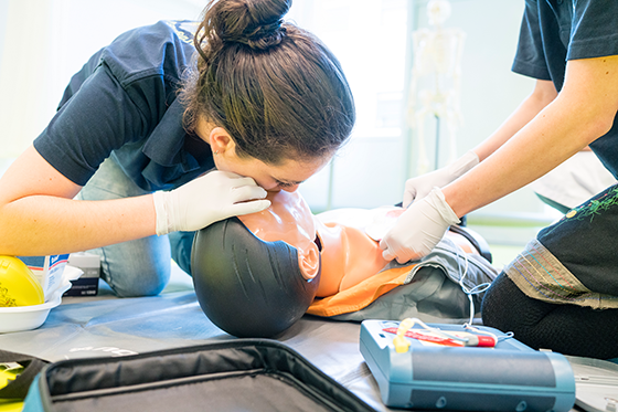 Twee mensen oefenen reanimatie op een trainingspop. Eén geeft mond-op-mondbeademing terwijl de ander een defibrillator voorbereidt.