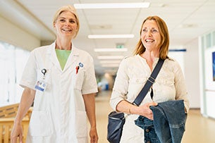 Een arts in een witte jas en een patiënt lopen glimlachend door de gang van het UMC Utrecht, waarbij de patiënt een jas en tas vasthoudt