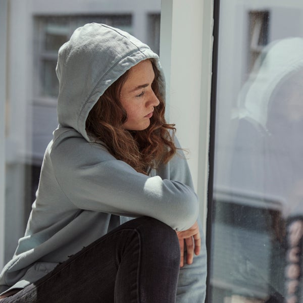 Een jonge vrouw in een grijze hoodie kijkt uit het raam.