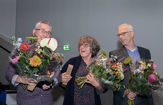 Drie opgetogen academici met boeketten bloemen vieren een onderscheiding tijdens een onderwijssymposium