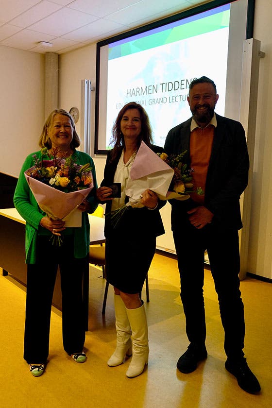 Drie personen staan glimlachend voor een scherm, waarbij twee van hen bloemen vasthouden, tijdens de Harmen Tiddens Educational Grand Lecture 2023