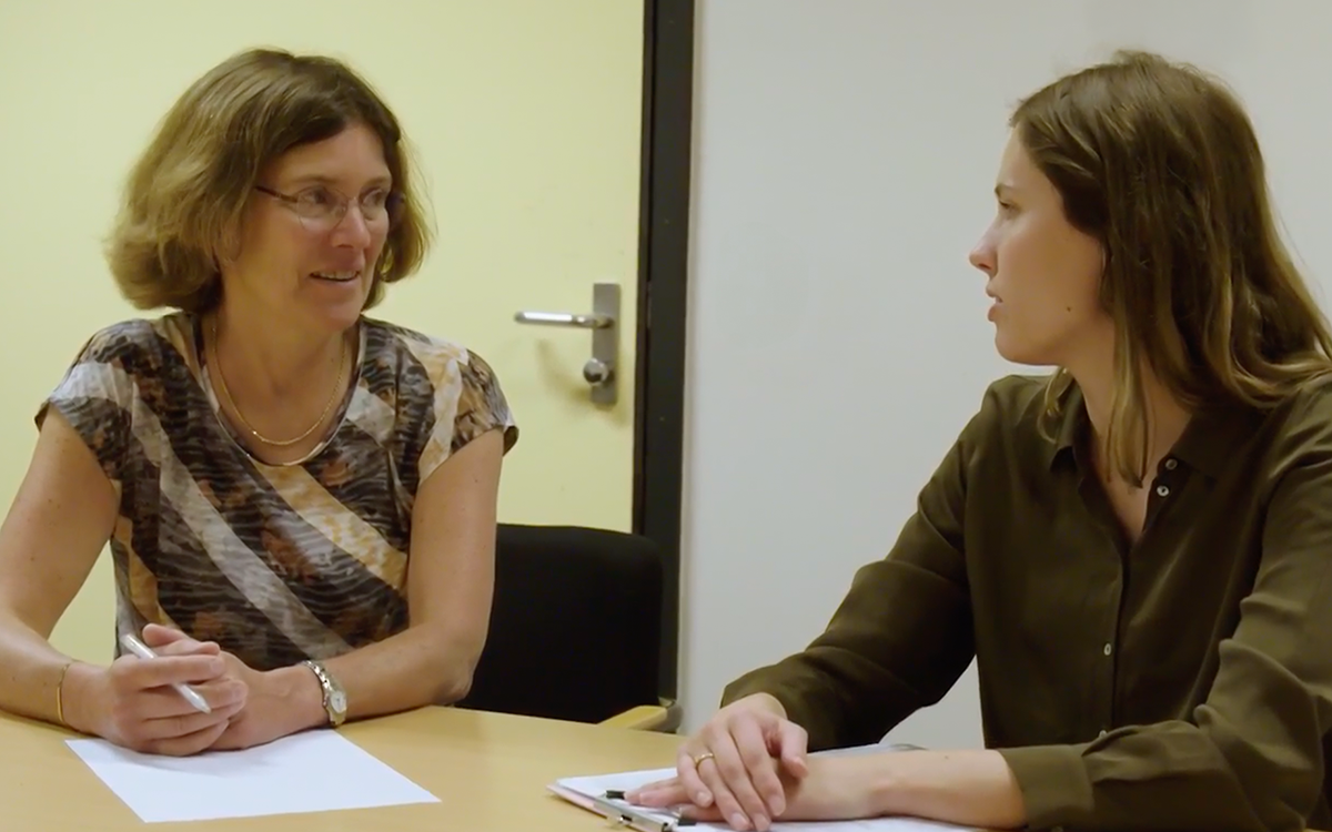 Een vrouw zittend aan een tafel met een vrouw die een pen vasthoudt in gesprek