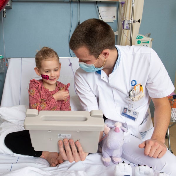 Meisje en zorgverlener zitten op een ziekenhuisbed en kijken naar een tablet.