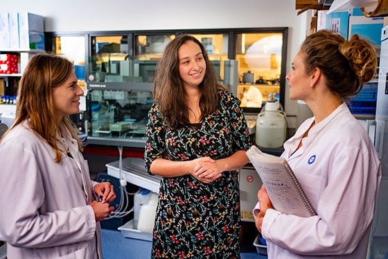 Drie vrouwelijke onderzoekers bespreken resultaten in een laboratoriumomgeving, waarvan twee een witte laboratoriumjas dragen en de derde in een jurk