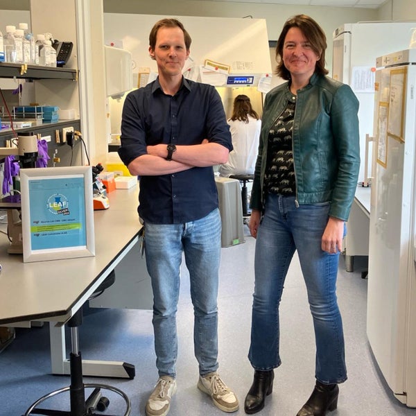 Twee onderzoekers staan in een laboratorium vol apparatuur en chemicaliën, glimlachend naar de camera. Een bord met "LEAF" is op de voorgrond te zien.