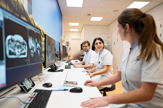Drie medische professionals in witte jassen werken samen aan computers, kijkend naar beeldschermen met medische scans, in een moderne kantooromgeving
