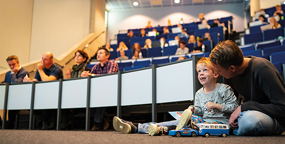 Een kind speelt met speelgoedauto's op de vloer van een collegezaal terwijl leerlingen op de achtergrond aan tafels zitten en luisteren.