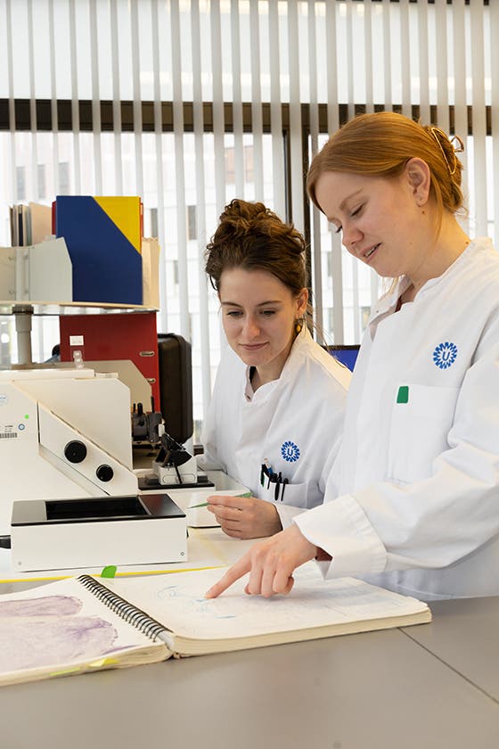Two female scientists in lab coats examining data and discussing over a notebook in a laboratory