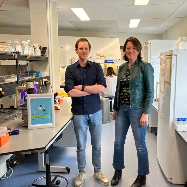Twee onderzoekers staan in een laboratorium vol apparatuur en chemicaliën, glimlachend naar de camera. Een bord met "LEAF" is op de voorgrond te zien.