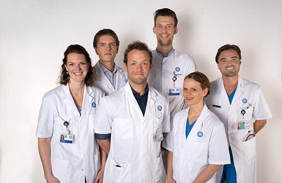 Zes medische professionals in witte jassen poseren voor de camera tegen een witte achtergrond, glimlachend en professioneel