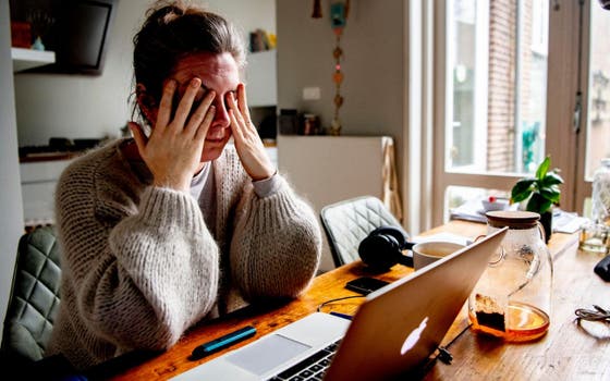 Een vermoeide vrouw met zit met haar handen voor haar ogen aan de keukentafel.