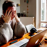 Een vermoeide vrouw met zit met haar handen voor haar ogen aan de keukentafel.